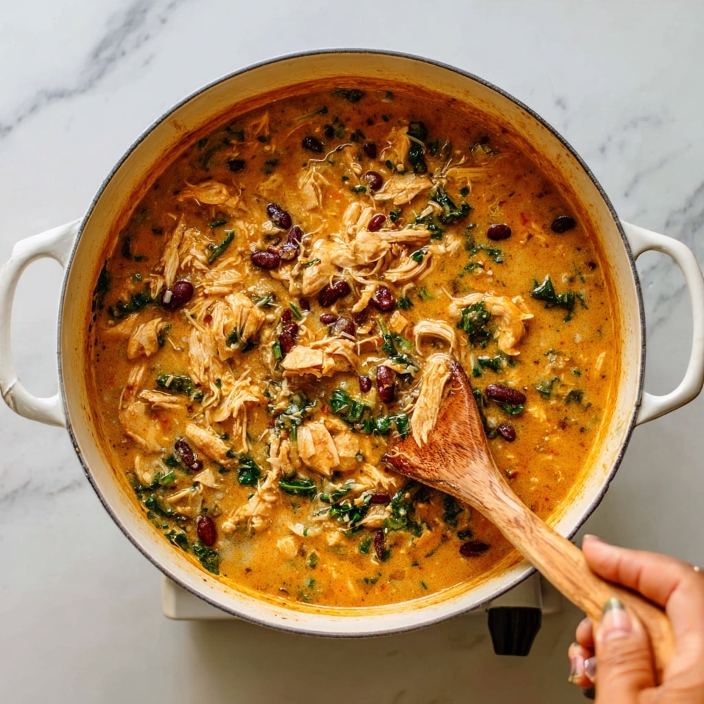 The image shows a white bowl filled with a creamy orange soup with visible chunks of shredded chicken, black beans, yellow corn, and green pepper pieces floating evenly throughout. On top, there are two bright green lime wedges placed slightly overlapping, next to a small bunch of fresh cilantro leaves adding a vibrant green contrast. A silver spoon rests inside the bowl on the left side, partly submerged in the soup. The bowl sits on a white marbled surface with faint gray veins and patterns, giving a clean and fresh background. photo taken with an iphone --ar 4:5 --v 7