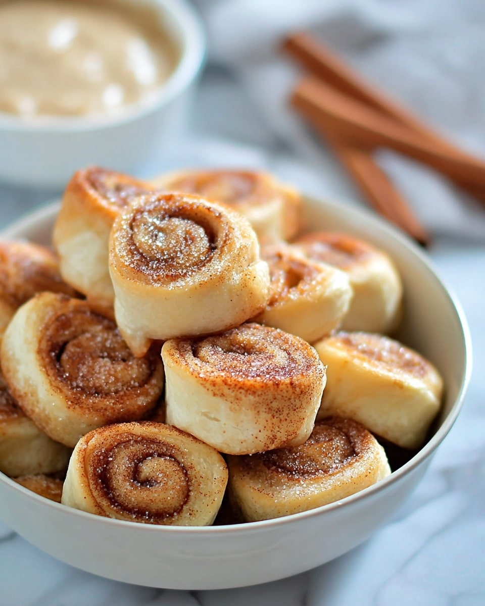 The image shows a white bowl filled with about twenty small cinnamon rolls arranged close together. Each cinnamon roll has a light golden-brown outer layer with a soft, doughy texture, and the inner swirled layer is a darker brown, covered lightly with a sprinkle of cinnamon sugar. Around the bowl, there are two whole cinnamon sticks placed on a white marbled surface. In the background, a white bowl with more cinnamon sugar and a few more cinnamon rolls stacked can be seen softly out of focus. Photo taken with an iphone --ar 4:5 --v 7