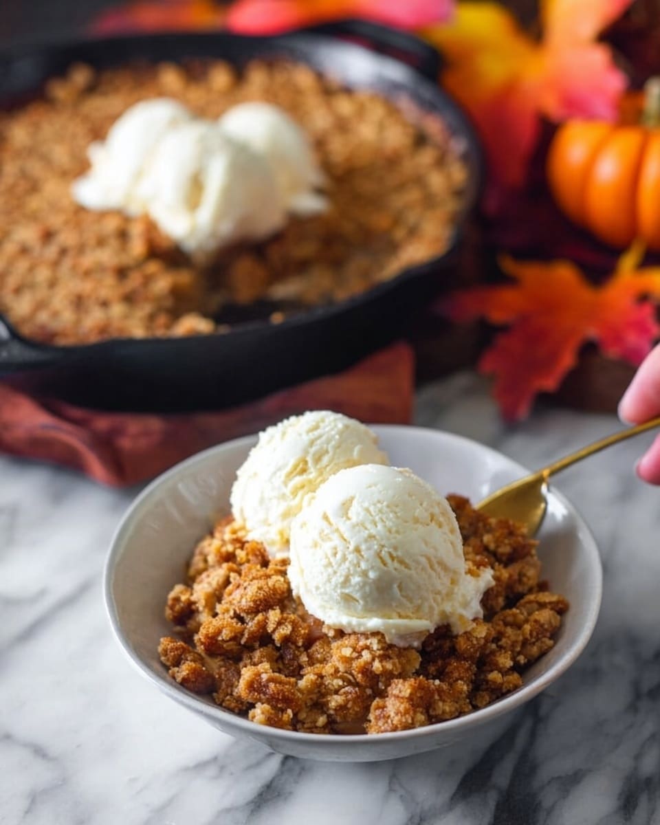 A white bowl is filled with one layer of crumbly brown pumpkin crumble topped with a single scoop of smooth white vanilla ice cream, held by a woman's hand. In the background, a black cast iron pan sits on a white marbled surface, containing a thick layer of the same crumbly pumpkin crumble topped with three scoops of vanilla ice cream. Around the pan, there are out-of-focus orange, red, and yellow autumn leaves that add warm colors to the image. The golden spoon rests inside the pan, slightly dipped into the crumble. Photo taken with an iphone --ar 4:5 --v 7