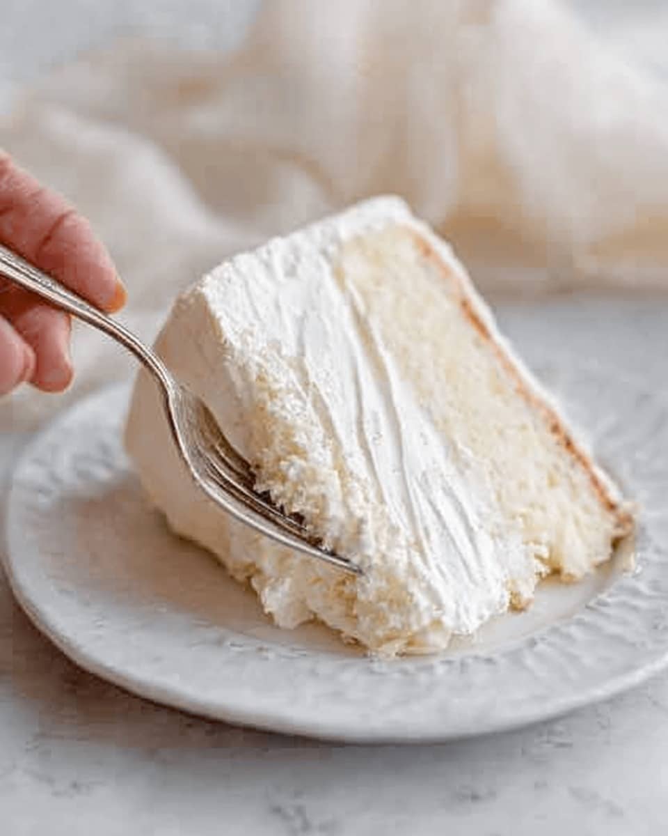 A two-layer cake sits on a clear glass cake stand. The cake is covered in smooth, creamy white frosting with soft swirling patterns. A large, delicate white flower decoration is placed on the side where the two cake layers meet. In the background, two tall white candles in metal holders are lit, adding a warm tone to the scene. The cake and accessories rest on a white marbled surface with white cloth napkins nearby, and a stack of white plates with silver forks is placed to the right. photo taken with an iphone --ar 4:5 --v 7