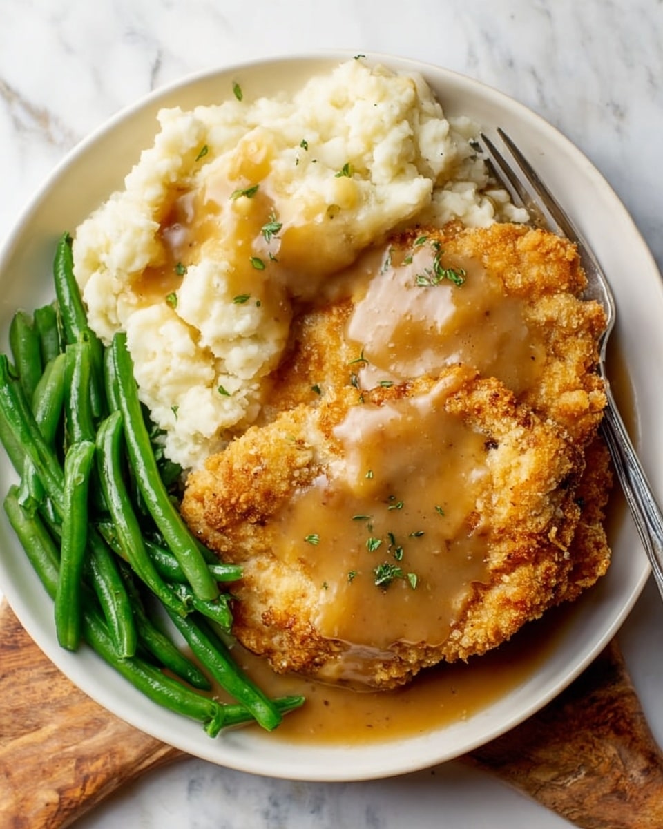 A white plate on a white marbled surface holds a comforting meal with three main layers: on the bottom, bright green cooked green beans arranged along the left side; next to them, creamy mashed potatoes in a large portion on the right side, topped with a layer of smooth brown gravy spreading across the surface; on top of both green beans and mashed potatoes are two large, golden-brown crispy fried pork chops covered partly by the same brown gravy. A silver fork rests on the right side in the mashed potatoes. Small green herb bits are sprinkled lightly on the dish photo taken with an iphone --ar 4:5 --v 7