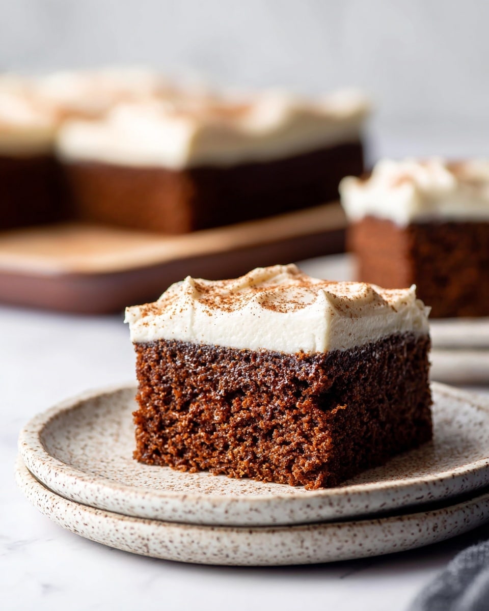 A square piece of chocolate cake is placed on a white plate with a decorative brown pattern along the edge. The cake has two layers: a thick, moist dark brown chocolate base and a thick white cream layer on top. On the cream, there is a swirl of whipped cream dusted with cocoa powder. A cinnamon stick lies next to the cake on the plate. The plate rests on a dark cloth over a white marbled surface, with soft background elements including a white cup and more whipped cream visible. Photo taken with an iphone --ar 4:5 --v 7