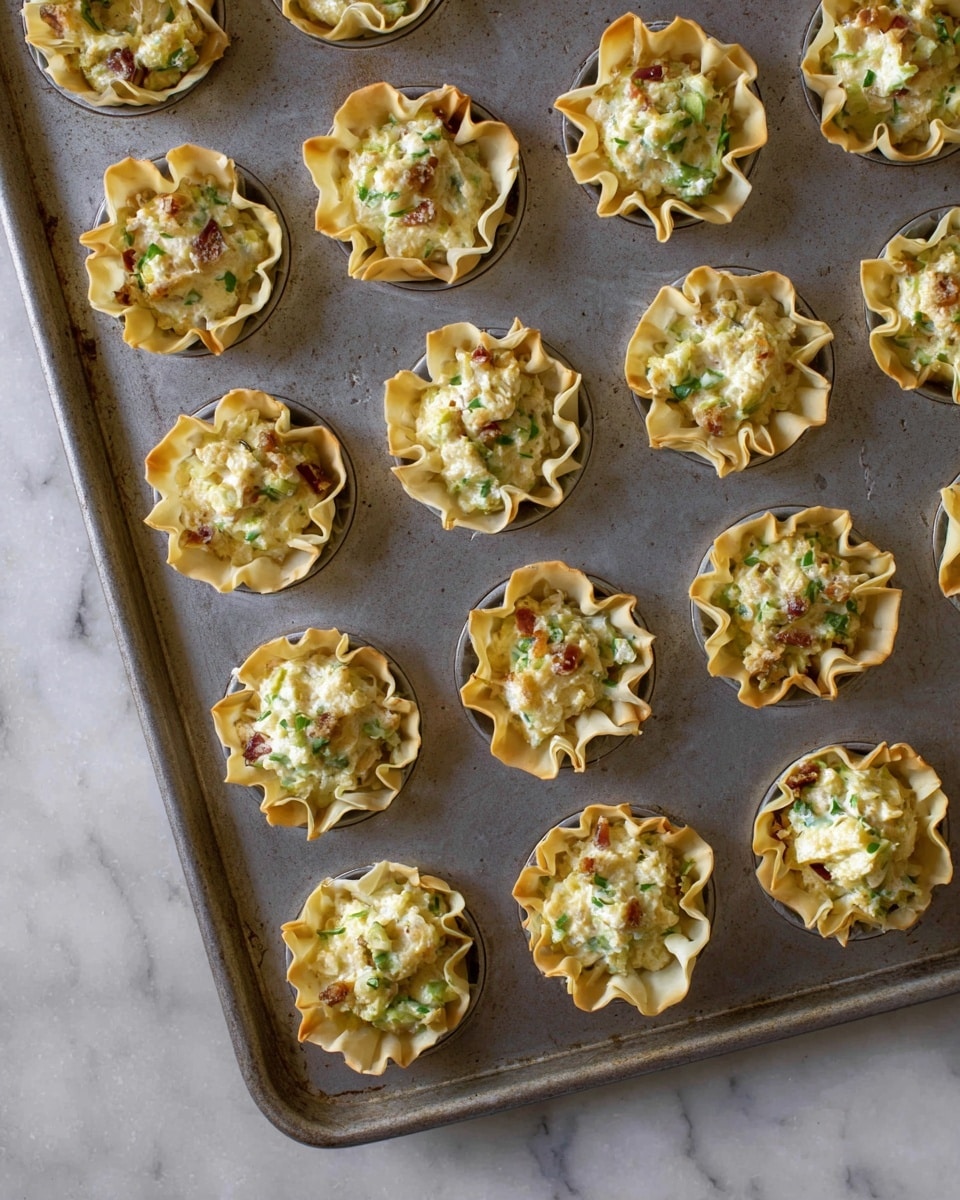 The image shows a baking tray filled with small phyllo pastry cups arranged in a neat grid. Each pastry cup has ruffled edges and is filled with a creamy mixture that looks soft and slightly chunky, with visible bits of green herbs and small pieces of browned ingredients. The pastry cups are light golden brown with a slightly crispy texture, and the filling has a pale yellow color mixed with flecks of green and caramelized brown. The tray sits on a surface with a white marbled texture. photo taken with an iphone --ar 4:5 --v 7