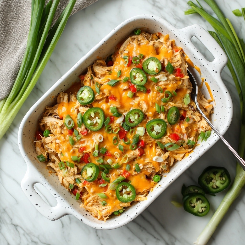 A close-up image of a bowl filled with shredded cooked chicken mixed with small pieces of diced red and green bell peppers, green peas, and finely chopped onions. The dish is topped with melted orange cheese sauce drizzled unevenly over the chicken and vegetables. Thin round slices of fresh green jalapeño peppers and chopped green onions are scattered on top, adding color and texture. The bowl is white with a black speckled pattern, and a silver fork rests inside it. In the background, out of focus, are some green onion stalks lying on a white marbled surface. photo taken with an iphone --ar 4:5 --v 7