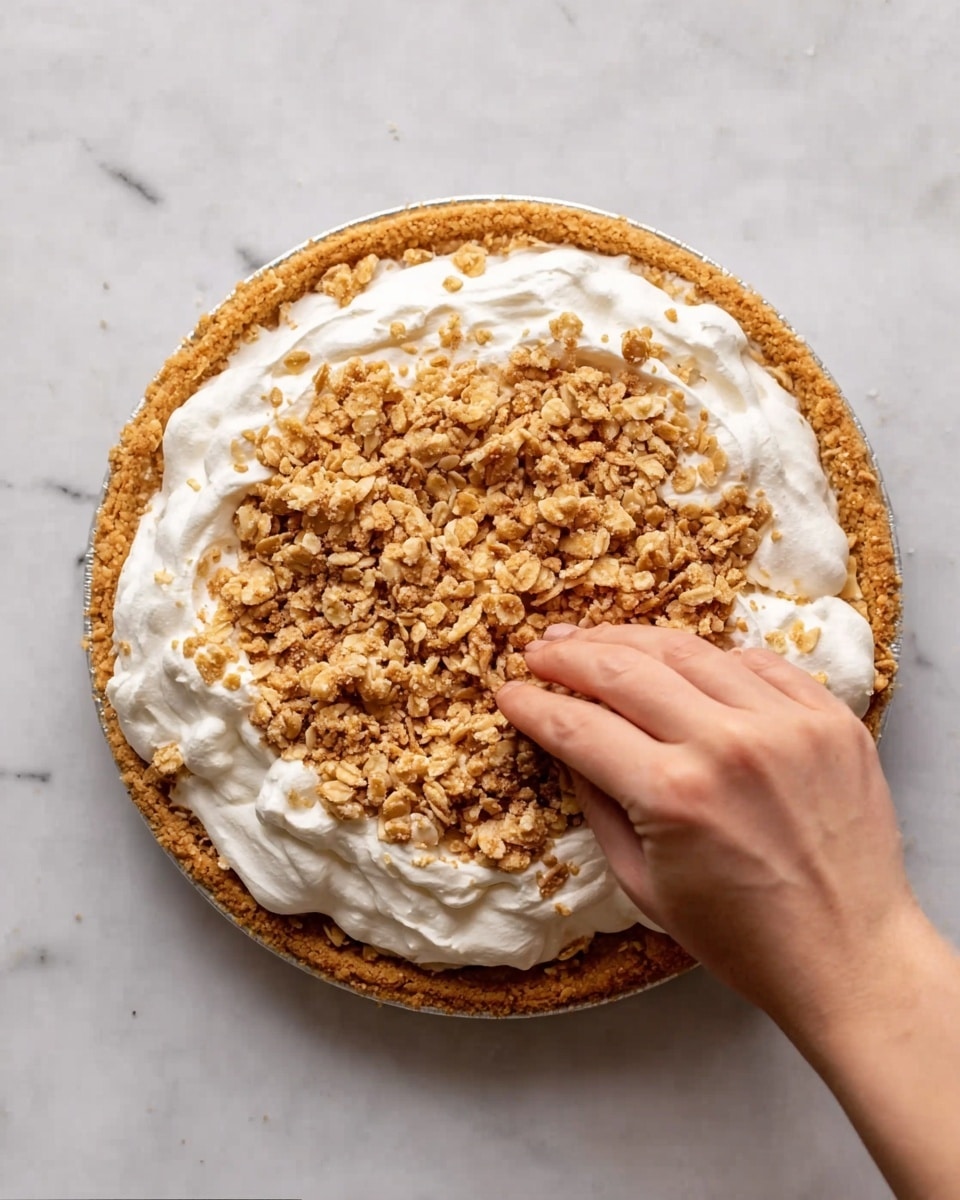 The image shows a clear glass pie dish with a crumbly golden brown crust at the bottom and along the sides. Inside the crust, there is a thick, white, smooth layer of whipped cream filling, topped with a generous layer of crumbled light brown cookies or biscuit pieces. Some crumbs are scattered around the pie dish on a white marbled surface with a folded dark blue cloth nearby. In the background, there is a white bottle with milk. The photo is taken with an iphone --ar 4:5 --v 7