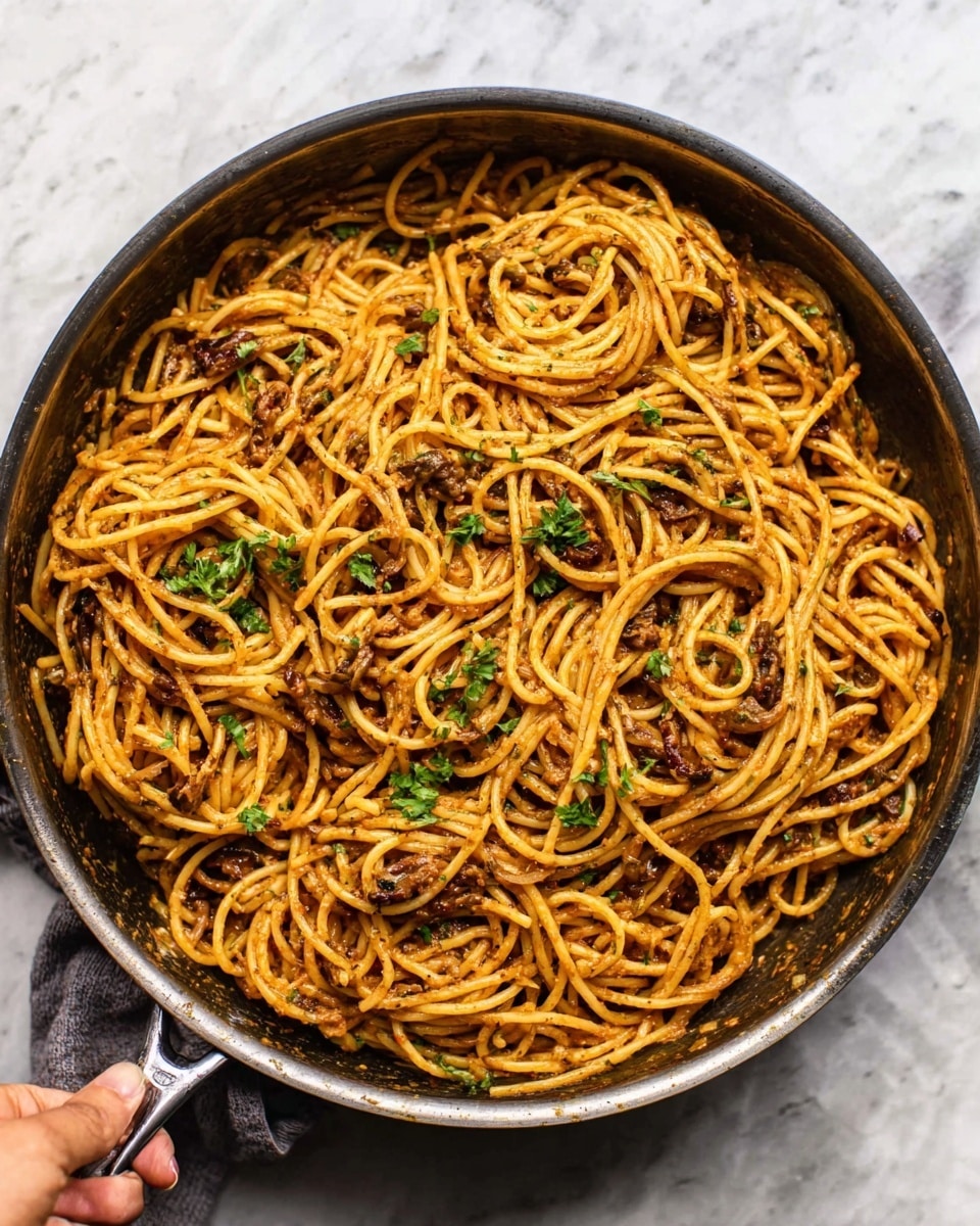 The image shows a bowl of spaghetti pasta with a rich, creamy sauce, light brown in color with small bits of herbs and spices spread evenly throughout. The spaghetti is fully coated, slightly shiny with a smooth texture. On top, a few fresh green herb leaves are scattered as garnish. One side of the bowl holds two pieces of toasted bread, golden brown with a crisp texture. The bowl is white, held by a woman's hand, and all of this is placed on a white marbled surface. photo taken with an iphone --ar 4:5 --v 7