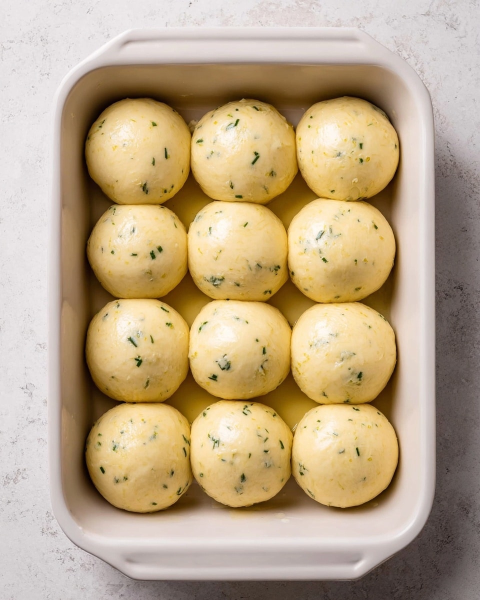 The image shows a close-up view of a group of soft dinner rolls arranged tightly together in a tray. The rolls have a shiny golden-brown top with a glowing glaze and are sprinkled with small bits of green herbs and white garlic pieces, adding texture and color contrast. The rolls appear fluffy and light inside, especially visible on one that is partially pulled apart, revealing a soft, white interior. The background features a white marbled texture that contrasts with the warm tones of the bread. photo taken with an iphone --ar 4:5 --v 7