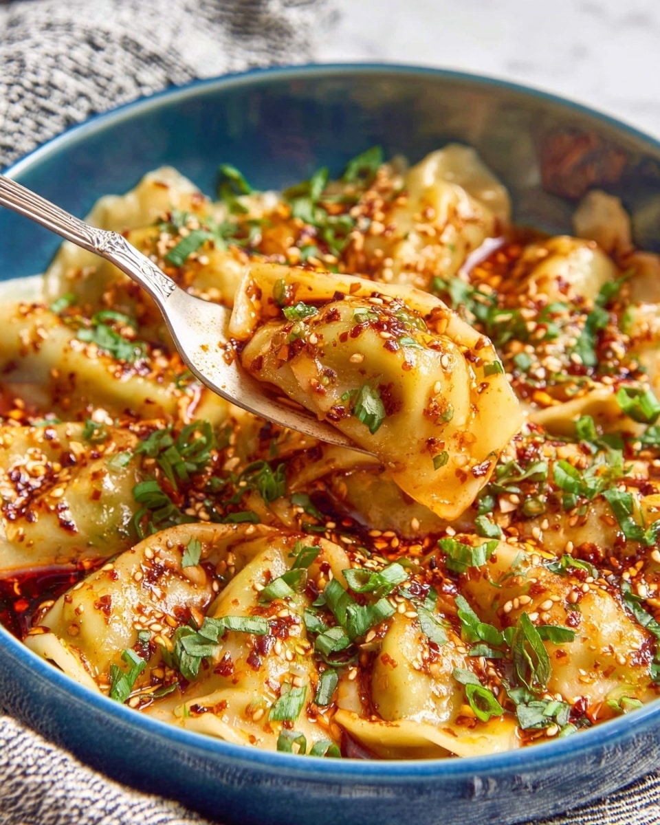 A close-up of a blue bowl filled with soft, golden dumplings covered in bright orange-red chili oil sauce with flecks of chili flakes and sesame seeds scattered on top. The dumplings have a smooth, slightly shiny texture and are garnished with chopped fresh green herbs and chopped scallions. A silver fork lifts one dumpling above the bowl, showing its curved, folded shape coated in the spicy sauce. The bowl sits on a white marbled surface with a textured striped cloth in the background. photo taken with an iphone --ar 4:5 --v 7
