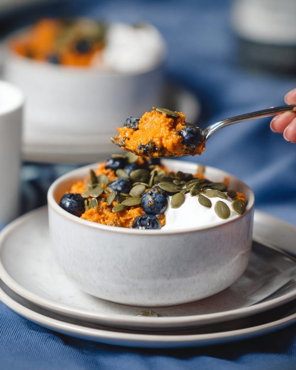The image shows a white bowl filled with a textured orange mixture topped with green pumpkin seeds and a dollop of white cream on the right side. There are dark blue blueberries scattered within and around the orange mixture inside the bowl. A silver spoon is lifting a portion of the orange mixture with some white cream and pumpkin seeds, held by a woman's hand. The bowl is placed on a stack of white plates with a white marbled surface beneath. In the blurred background, another white bowl with similar contents is partially visible. photo taken with an iphone --ar 4:5 --v 7