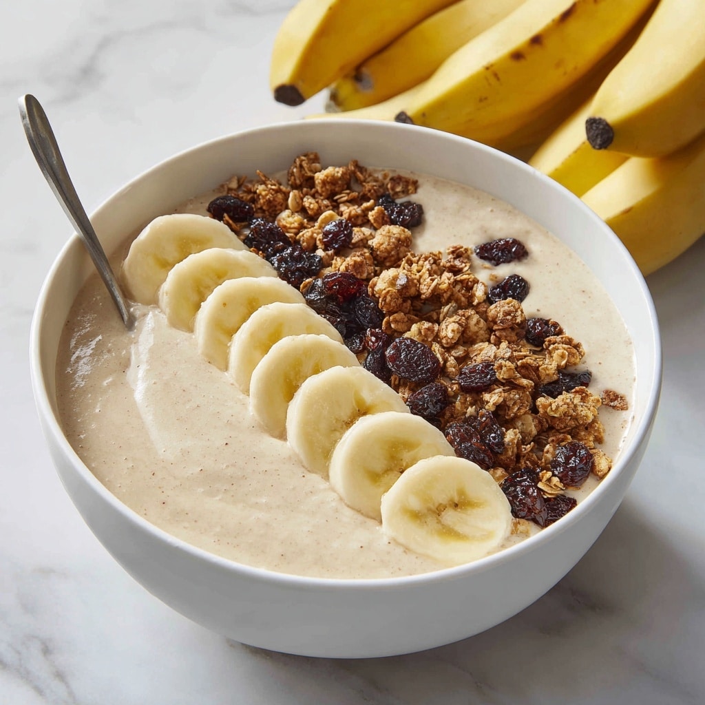 A white bowl filled with a thick, creamy beige smoothie base with a smooth texture covers most of the bowl's surface. On top, there are three layers arranged in a neat row along one side: the first layer consists of light yellow banana slices with a soft, slightly shiny texture; next to it is a layer of dark brown granola clusters with bits of dried raisins, giving a rough and crunchy feel; scattered slightly over both the smoothie and toppings are a few more granola pieces. A silver spoon rests on the left side of the bowl, partially dipped into the smoothie. In the background, a bunch of small ripe bananas sit on a white marbled textured surface. Photo taken with an iphone --ar 4:5 --v 7