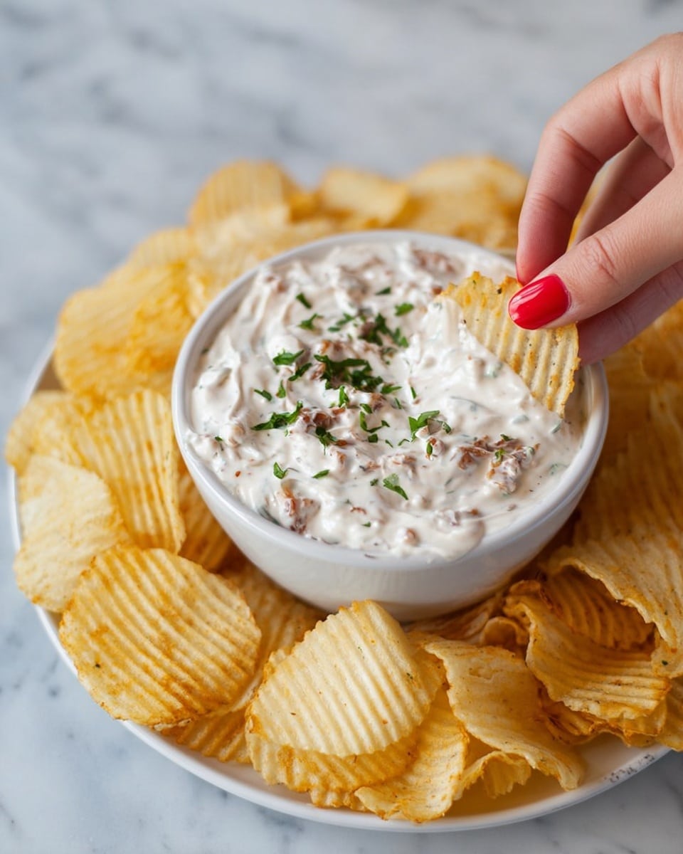 A white bowl filled with a creamy, thick white dip that has small brown bits mixed inside, topped with chopped green herbs. The bowl sits on a white plate surrounded by many golden ridged potato chips. A woman's hand with red nail polish is dipping one ridged chip into the creamy dip. The background shows a white marbled texture. photo taken with an iphone --ar 4:5 --v 7