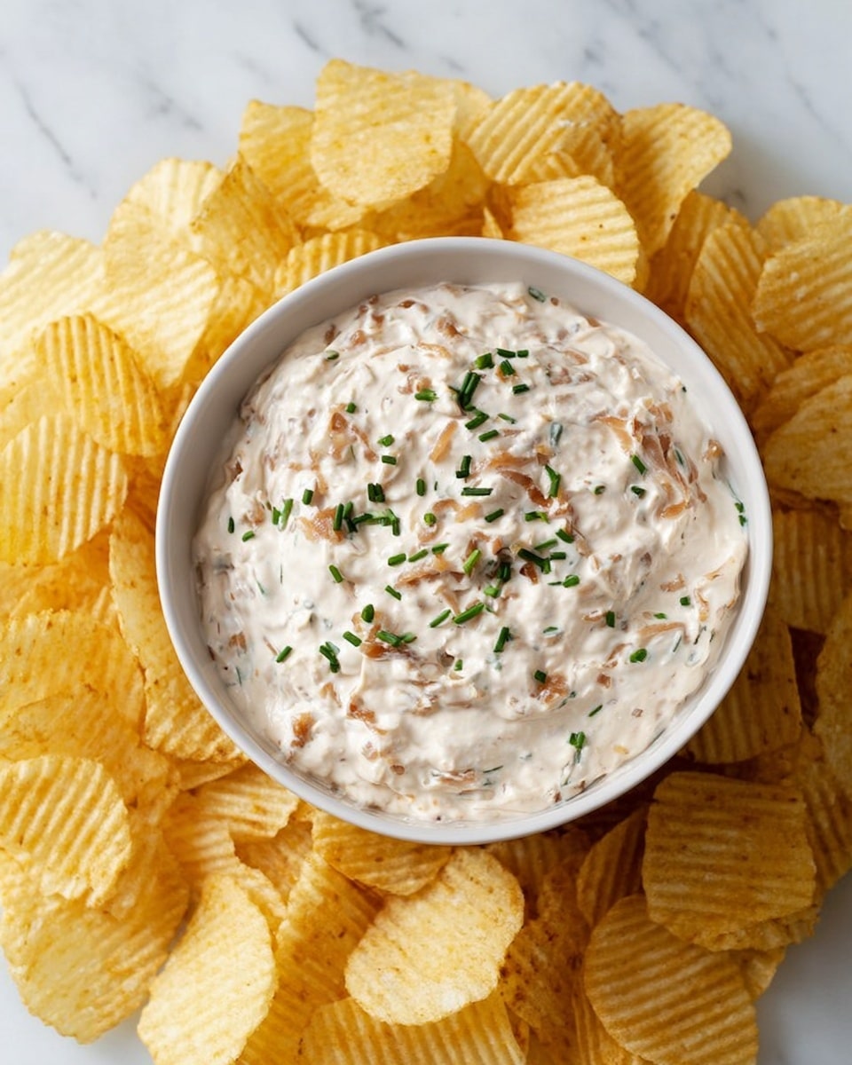 A white bowl filled with creamy, thick dip that has small bits of caramelized onion mixed throughout, topped with chopped green herbs. The bowl is placed in the center of a pile of ridged, golden-yellow potato chips spread evenly around it. The whole setup sits on a white marbled surface, creating a clean and fresh look. photo taken with an iphone --ar 4:5 --v 7