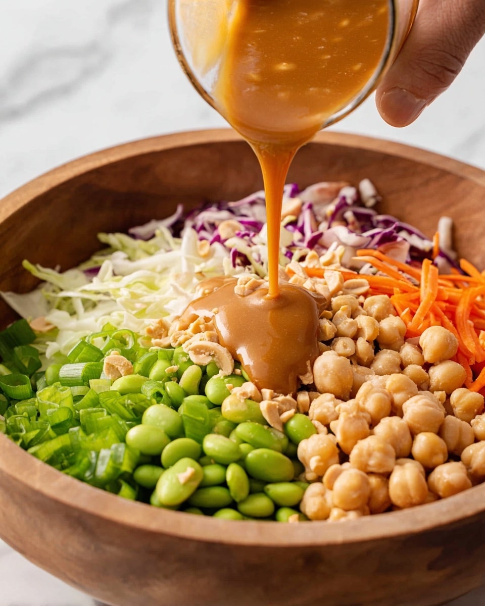 A close-up image shows a wooden bowl filled with fresh chopped green onions at the bottom left, bright green edamame beans in the middle top, beige chickpeas on the top right, chopped peanuts on the bottom right, and shredded white and purple cabbage with small pieces of orange carrot on the top left. Above these layers, a thick caramel-colored sauce is being poured from a small glass container held by a woman's hand, dripping fluidly onto the toppings. The background features a white marbled texture. Photo taken with an iphone --ar 4:5 --v 7