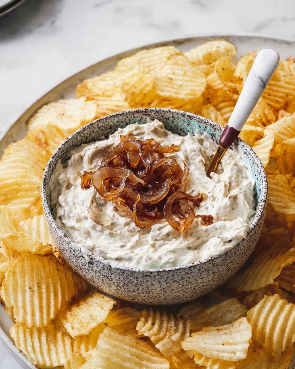 A rustic bowl with a speckled grey and white texture holds a creamy dip with a thick, smooth texture, topped with golden-brown caramelized onion slices that add a rich contrast. Inside the bowl, a white spoon with a brown-striped handle is partially dipped in the dip, resting on one side. Surrounding the bowl is a large round white plate filled with crispy, wavy potato chips that are light golden-yellow in color and have a crunchy texture. The whole setup is placed on a white marbled surface, creating a clean and bright background for the snack. photo taken with an iphone --ar 4:5 --v 7