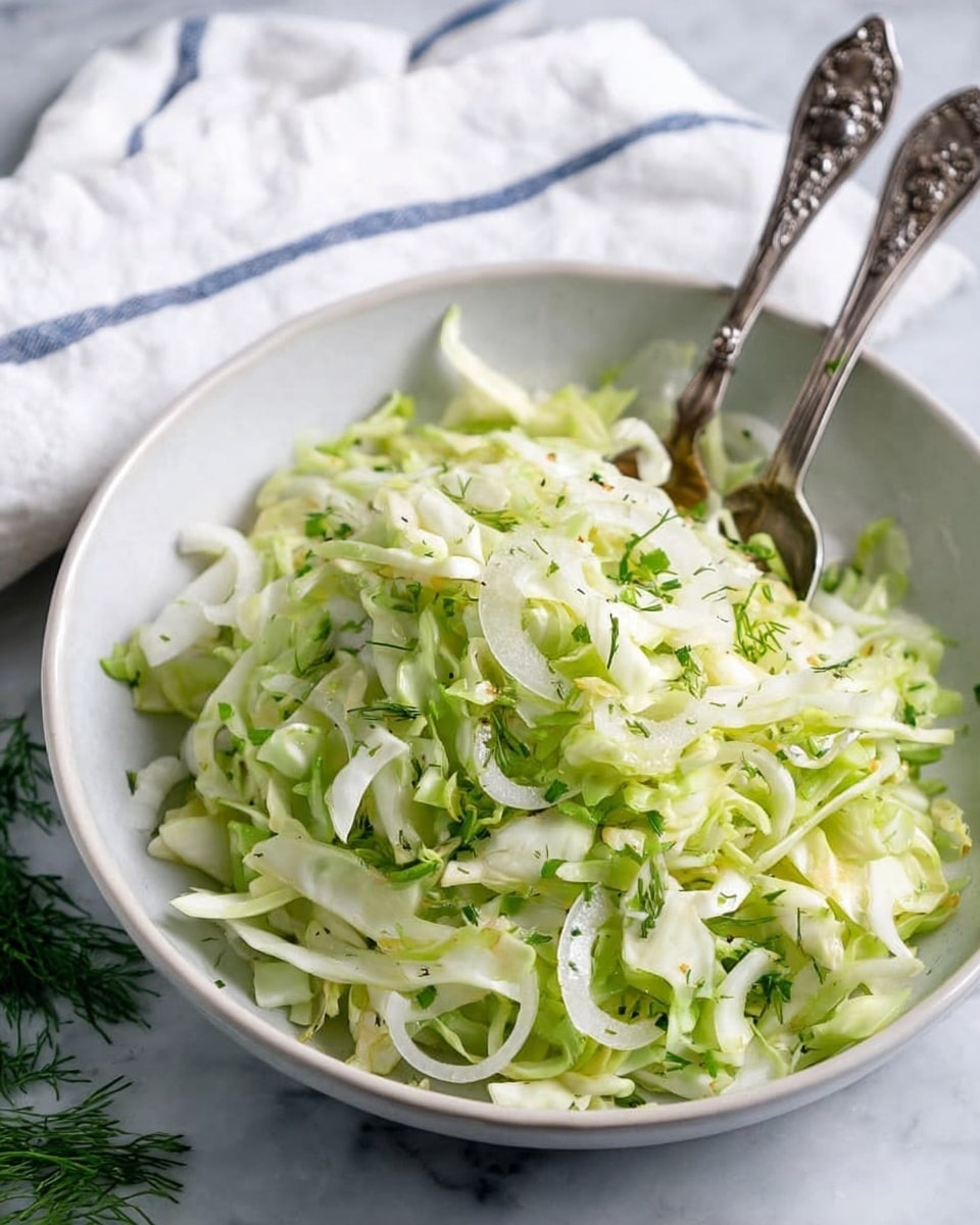 A white bowl filled with a fresh salad made of thinly sliced light green cabbage, white onion rings, and small pieces of green herbs sprinkled over the top. Two ornate silver spoons stand inside the bowl, slightly angled outward. The bowl sits on a white marbled surface with some green herb sprigs around it, and there is a white cloth with blue lines partially visible behind the bowl. The overall look is crisp and fresh. photo taken with an iphone --ar 4:5 --v 7
