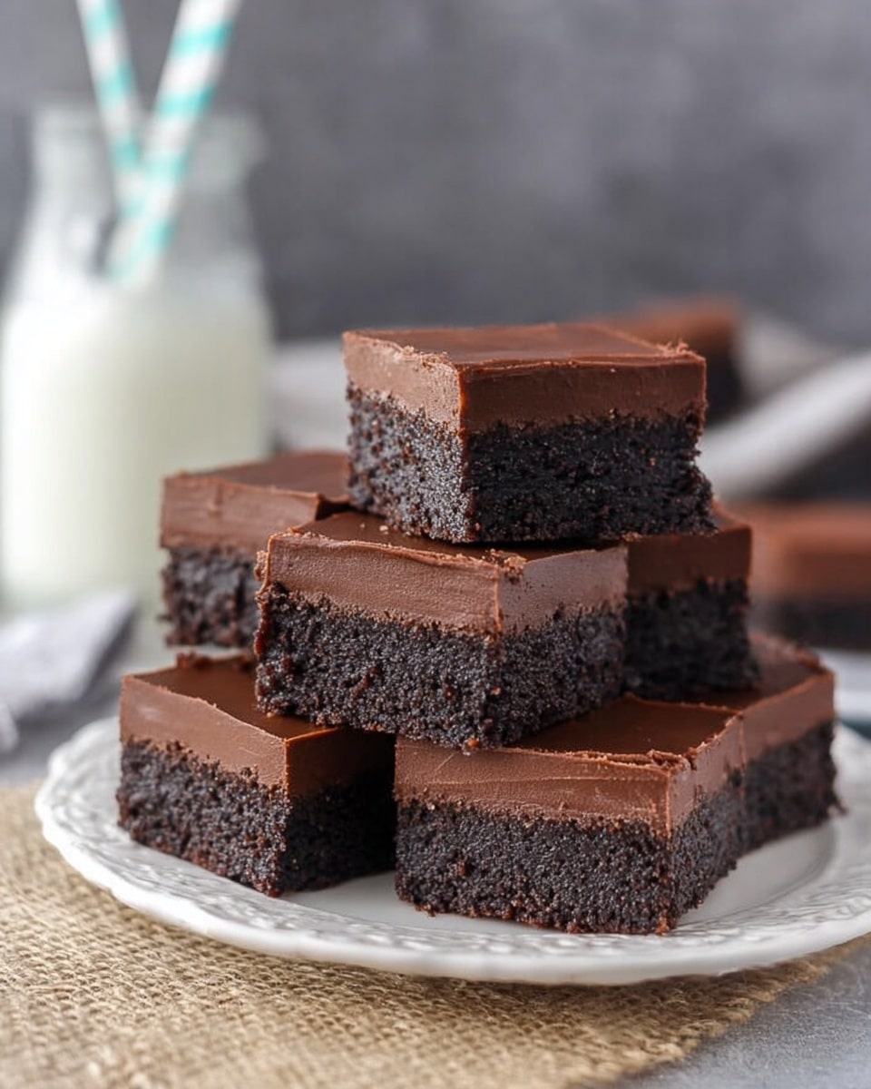 The image shows a stack of five square chocolate bars arranged on a white plate with a delicate embossed pattern. Each bar has two layers: a thick, dark, crumbly-looking bottom layer similar to dense cake or brownie, and a rich, smooth, dark chocolate top layer with a creamy texture. The bars are neatly cut into perfect squares, with the top bar slightly angled, revealing a creamy bite on one corner. The plate sits on a rough beige fabric, and behind it, there is a cool gray background and a blurred glass bottle filled with milk and a blue-and-white striped straw. Photo taken with an iphone --ar 4:5 --v 7