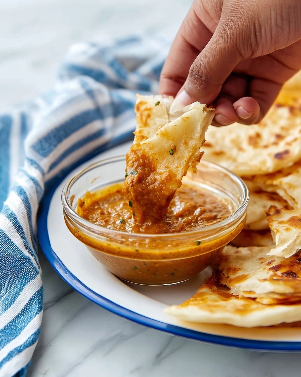 A white plate sits on a white marbled surface with a blue and white striped cloth under the plate. On the plate, there are five pieces of golden brown, flaky, folded flatbread arranged in a semi-circle. Each flatbread piece shows multiple thin layers with crispy, browned spots on the surface, giving a textured, slightly bubbled look. Next to the flatbreads is a white bowl filled with smooth orange curry topped with a swirl of white cream and small green chopped herbs, creating a colorful contrast. Photo taken with an iphone --ar 4:5 --v 7
