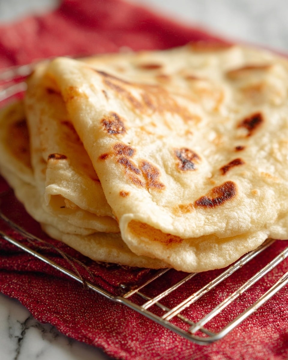 The image shows a close-up of two soft flatbreads stacked on top of each other, placed on a metal cooling rack over a red cloth with a subtle texture. The top flatbread is folded over, revealing a light golden-brown surface with darker toasted spots and a slightly powdery texture. The edges are soft and slightly uneven, showcasing the handmade look. The overall colors are warm beige and brown tones, with the cool metal rack and red cloth underneath adding contrast. The background is a white marbled texture. photo taken with an iphone --ar 4:5 --v 7