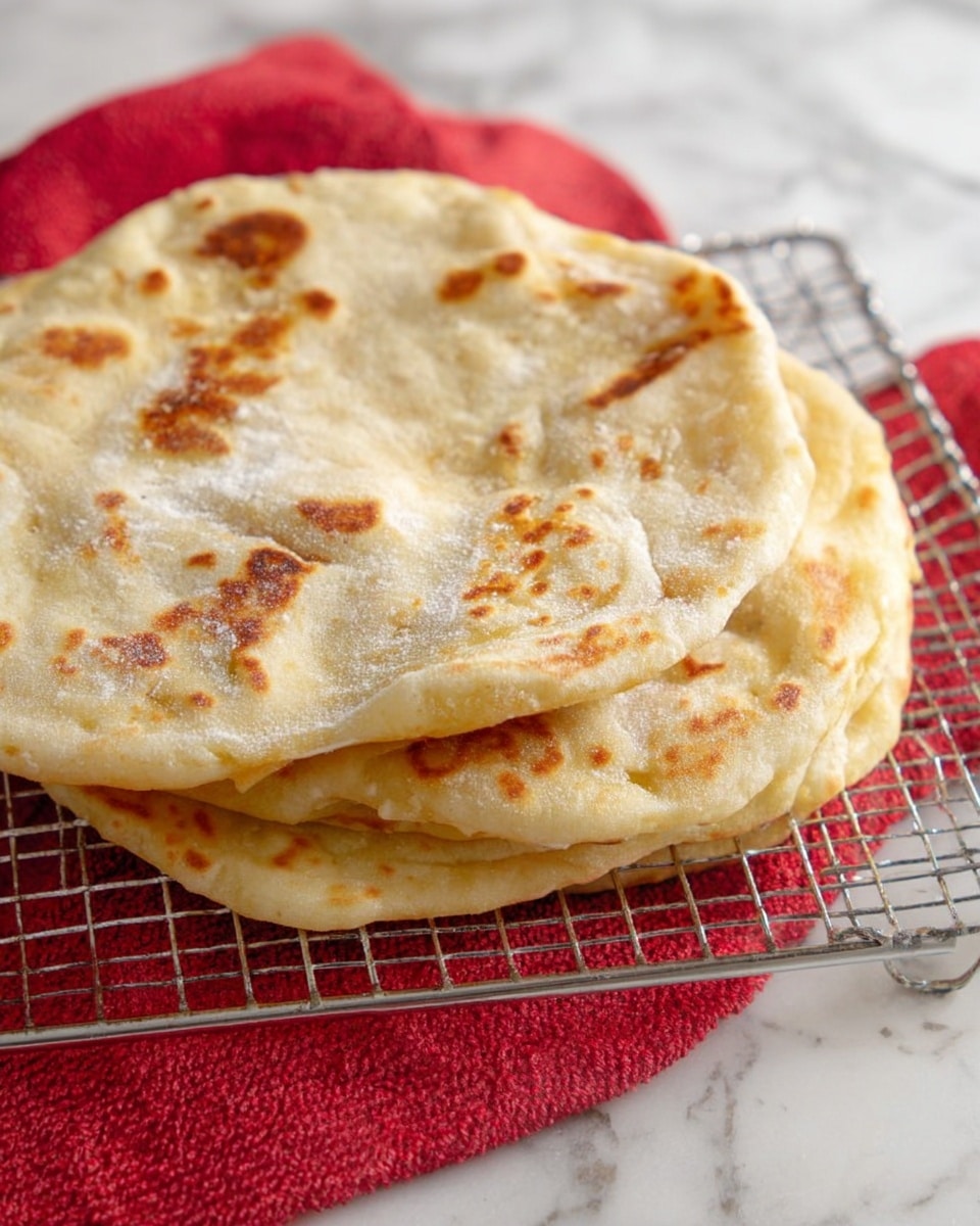 A stack of three round, flat breads with a light golden-brown color and some darker brown spots, showing a soft, slightly puffy texture with subtle dusting of flour on top, resting on a metal cooling rack placed over a red cloth, all set on a white marbled surface. photo taken with an iphone --ar 4:5 --v 7