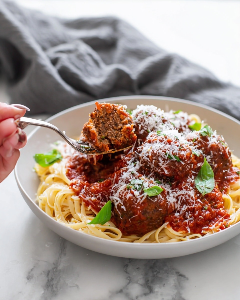 A white bowl filled with a base layer of long, thin, pale yellow spaghetti noodles. On top, there are four large brown meatballs covered with rich, chunky red tomato sauce. The sauce glistens and is thickly spread over the spaghetti and meatballs. Fresh green basil leaves are scattered around the plate, adding bright color. White grated cheese is sprinkled generously over the meatballs and sauce, giving a snowy texture on top. A woman's hand is holding a fork that slightly cuts into one meatball, showing its dense, crumbly brown inside. The background is a white marbled surface with a blurred grey cloth. Photo taken with an iphone --ar 4:5 --v 7