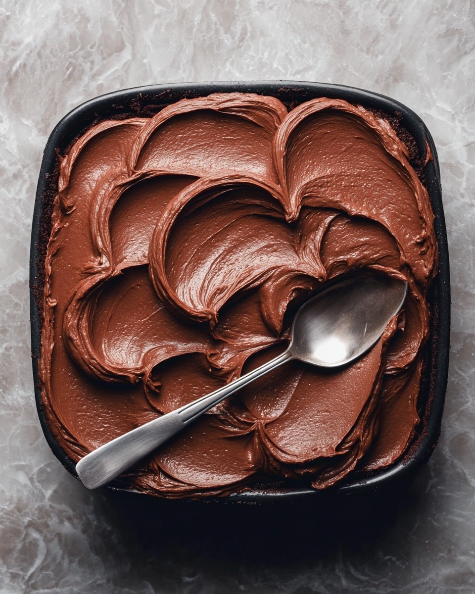This image shows a clear glass bowl filled with thick, glossy, dark brown chocolate batter. The batter has swirled and smooth textures, with shiny highlights reflecting the light. A white mixer paddle partially covered with the chocolate batter is positioned resting inside the bowl on the left side. The bowl sits on a white marbled surface. photo taken with an iphone --ar 4:5 --v 7