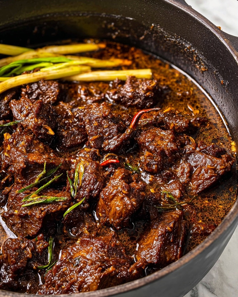 The image shows a close-up of a thick stew with chunky pieces of dark brown tender meat in a deep black pot. The meat is coated in a rich, glossy sauce with a slightly oily texture, visible bits of spices, and herbs. Several pale yellow stalks of lemongrass lay on top of the meat near the back left edge, adding contrast to the dark tones of the stew. The pot sits on a white marbled surface, enhancing the deep colors of the dish. photo taken with an iphone --ar 4:5 --v 7
