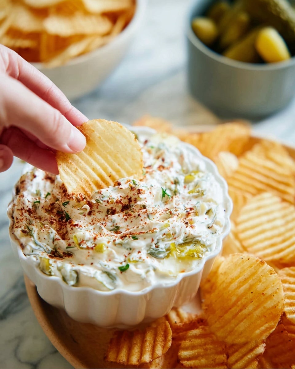 A close-up image showing a woman's hand holding a light yellow ridged potato chip dipped in a creamy white dip with visible green herbs and small yellow pickle pieces, topped with a sprinkle of red seasoning. The dip is in a white scalloped bowl filled almost to the top, with the creamy mixture textured with green herbs and diced pickles, and a light dusting of red spices on the surface. Around the bowl, there are more ridged chips, some inside a white bowl filled with golden yellow chips that have a slightly crisp texture. Part of a gray bowl with greenish pickles is visible at the top right. The setting features a white marbled textured surface. Photo taken with an iphone --ar 4:5 --v 7