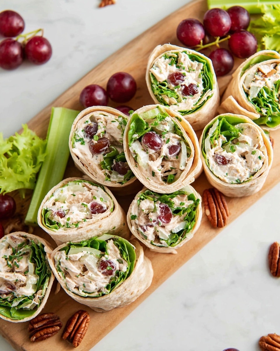 The image shows several wrap pinwheels placed on a wooden board. Each wrap has three main visible layers: the outer soft, light beige tortilla, a fresh green leafy lettuce layer inside, and a creamy white chicken salad filling mixed with small pieces of celery, red grapes, and bits of pecan nuts. The wraps are cut into thick, round slices and are garnished with small bits of green herbs on top, giving a fresh contrast. Around the board are scattered whole red grapes, celery stalks, and pecan pieces on a white marbled surface. Photo taken with an iphone --ar 4:5 --v 7