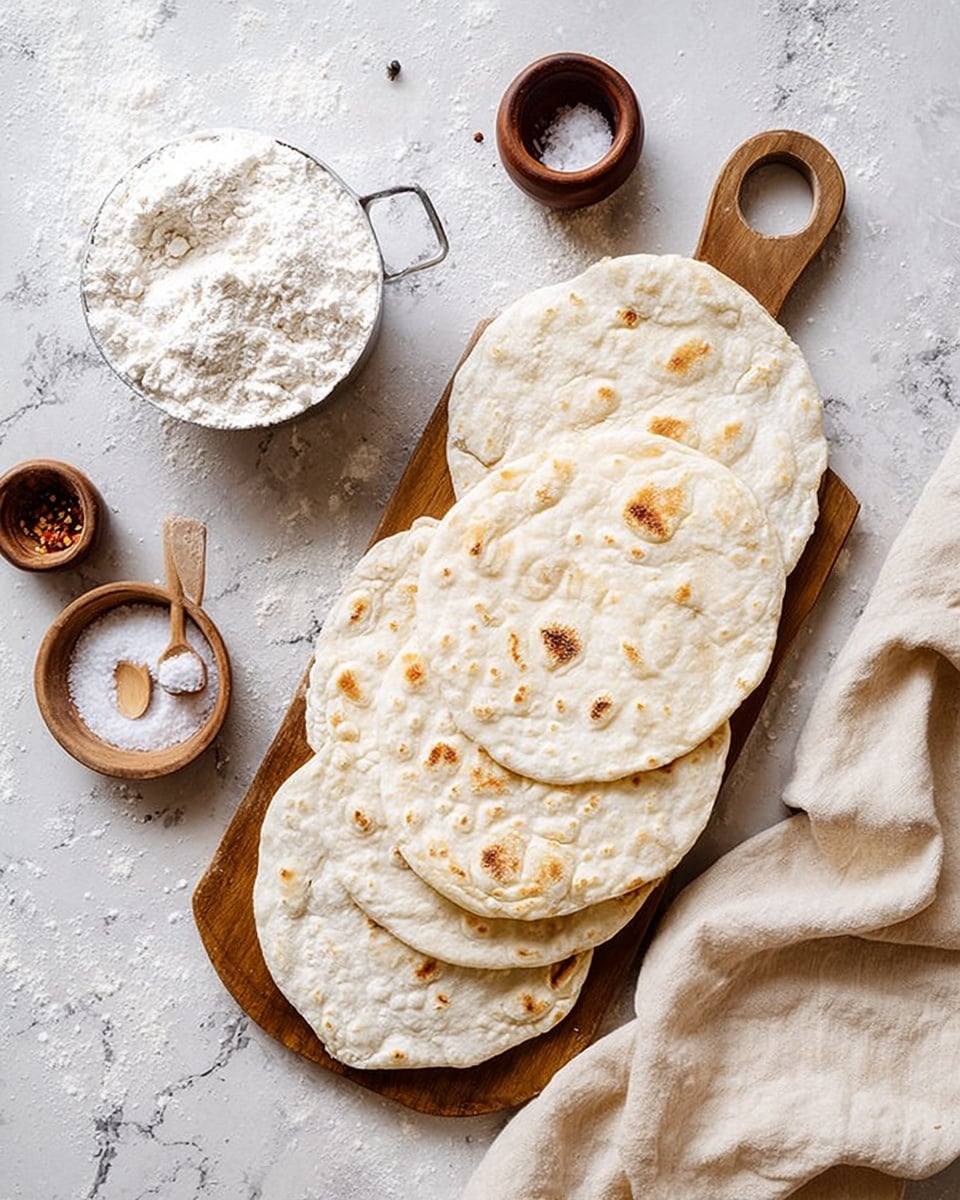 A wooden board is covered with six round, flat pieces of white bread with a lightly uneven surface and small brown spots from cooking, each layered slightly overlapping. To the left of the board, a metal measuring cup filled with white flour is placed on a white marbled surface, with some flour spilled around it. Nearby, there are four small brown wooden spice containers scattered and a small white bowl containing salt with a tiny wooden spoon. A light beige cloth covers the lower right corner of the image. The overall setting is bright with soft natural light. photo taken with an iphone --ar 4:5 --v 7