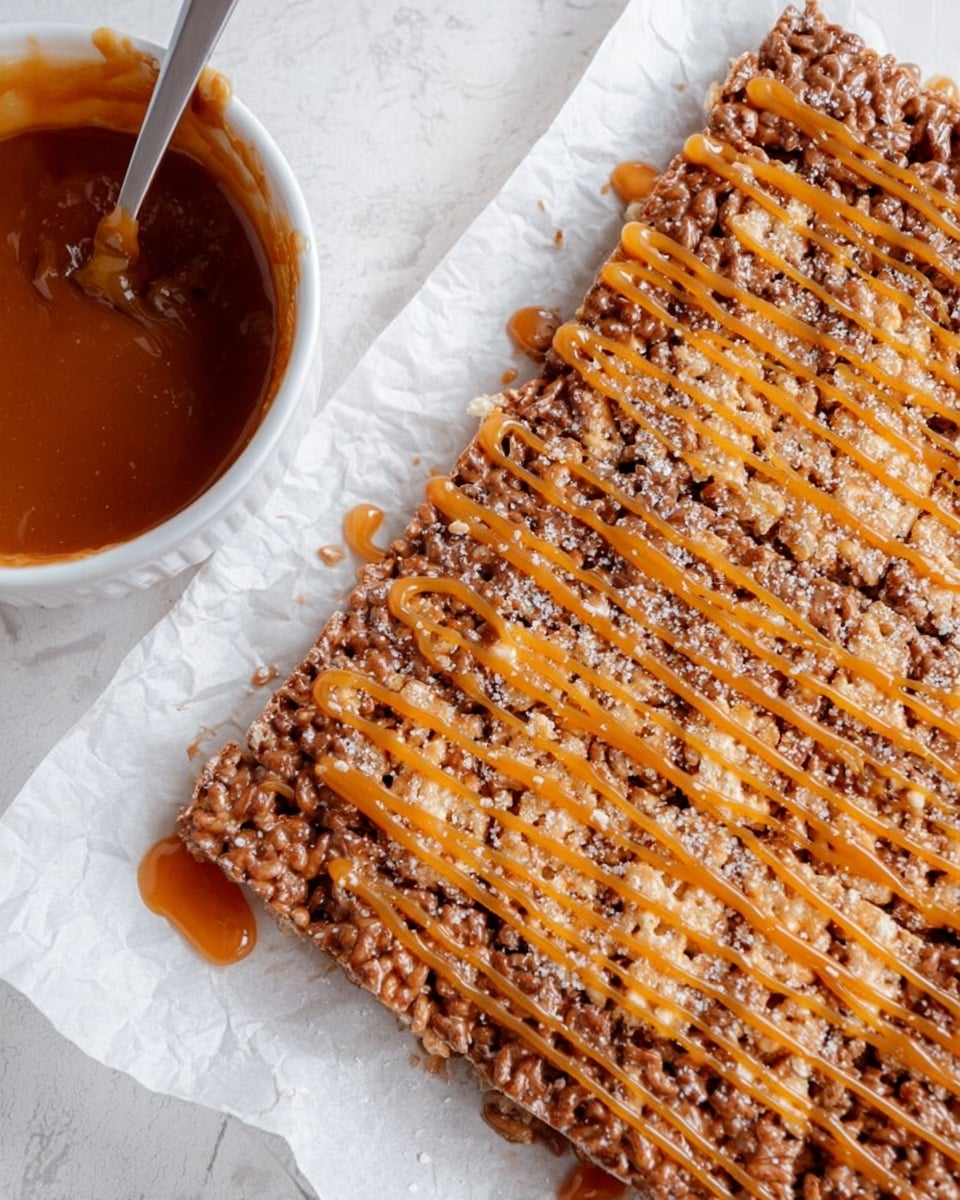 A close-up view of a large, rectangular crispy treat made from puffed rice and chocolate, drizzled in thin, uneven lines of orange caramel sauce across the top. The treat has a rough, textured surface with visible grains and a light sprinkle of finely crushed nuts or sugar crystals adding extra texture. It sits on a white sheet of baking paper, placed on a white marbled surface. To the left, there is a white glass bowl containing some melted caramel sauce, with a fork resting inside it, coated in the same sauce. photo taken with an iphone --ar 4:5 --v 7