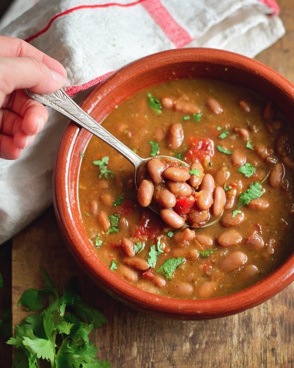 A rustic bowl filled with thick, brown bean soup with visible whole beans and chunks of red tomato throughout, garnished with small bright green cilantro leaves scattered on top. A woman's hand is holding a silver spoon scooping some of the soup from the bowl. The bowl is terracotta colored and sits on a wooden surface, with a white cloth featuring red stripes partially visible in the background and a bit of fresh green cilantro off to the side. The textures of the beans and broth look hearty and comforting. photo taken with an iphone --ar 4:5 --v 7