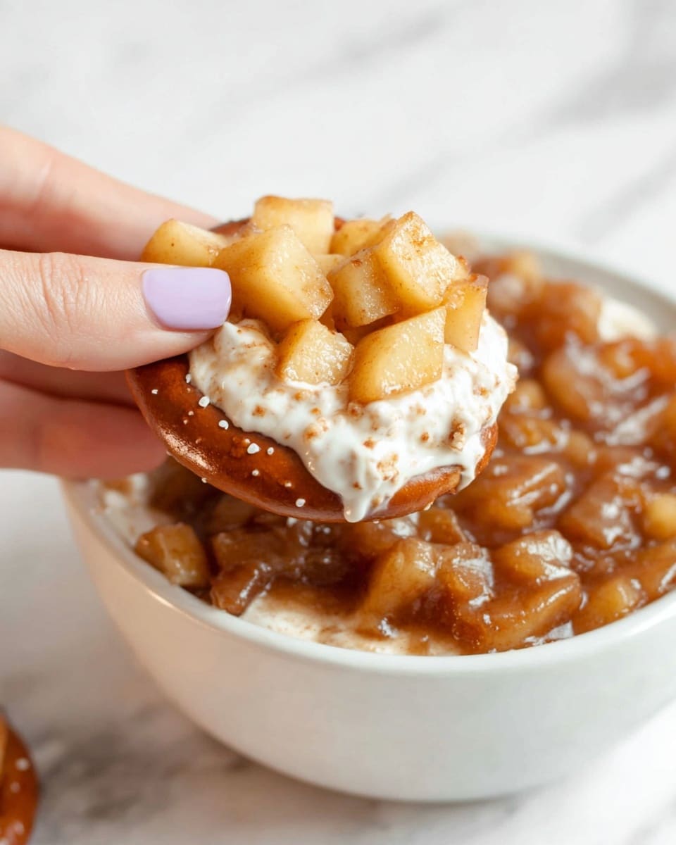 A white bowl filled with a layer of creamy white base topped thickly with a chunky, glossy brown mixture of diced apples and sauce. On the right side of the bowl, several small shiny pretzels rest on top of the apple layer. Around the bowl, on a wooden board, a few crunchy pretzels are scattered. In the background, there is a small white bowl also filled with the chunky apple mixture and a single pretzel on top, placed on a soft white cloth on a white marbled surface. photo taken with an iphone --ar 4:5 --v 7