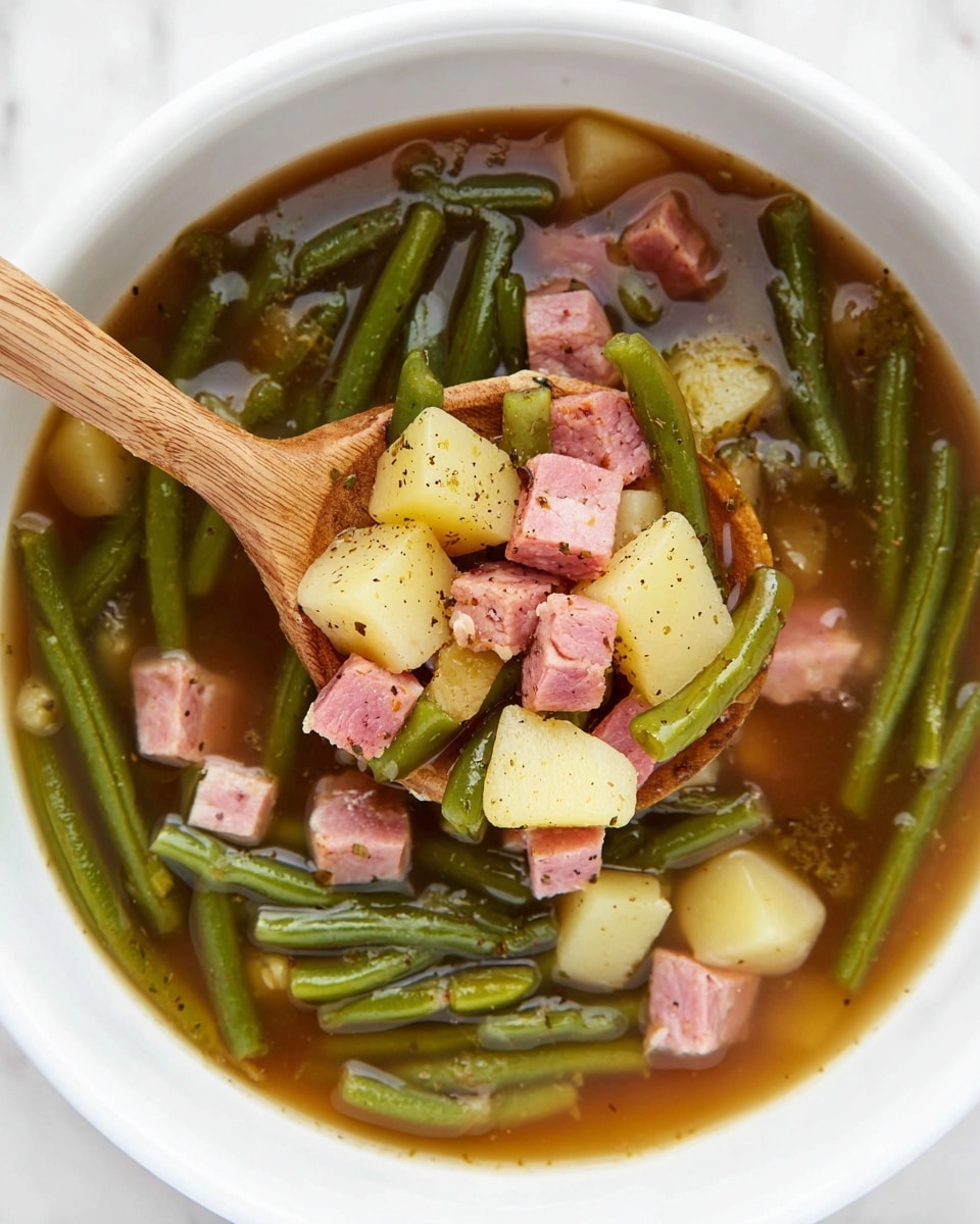 A white bowl filled with a clear brown broth containing bright green beans that form the first layer around the bowl, next a layer of small pale yellow potato cubes mixed with pinkish chunks of ham sprinkled with black pepper, all floating gently in the soup. A wooden spoon is partially submerged in the center of the bowl lifting a mix of green beans, potatoes, and ham cubes. The scene is set on a white marbled surface. photo taken with an iphone --ar 4:5 --v 7