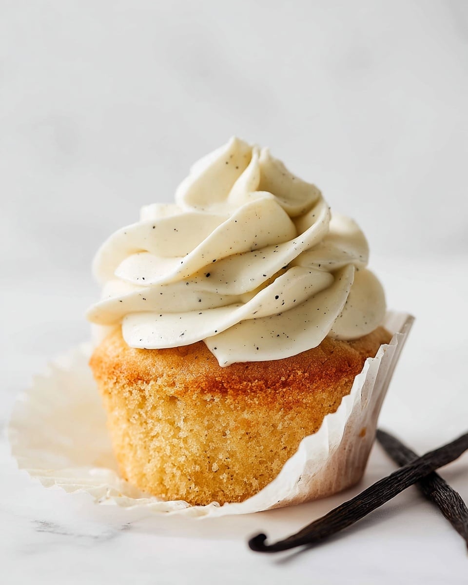 A white bowl filled with thick, swirled white frosting sits on a beige cloth with black grid lines, next to two cupcakes topped with one layer each of smooth, rose-shaped white frosting. To the left, a clear piping bag filled with the same white frosting is placed near metal beaters covered with frosting, while three yellow-white candy melts are scattered to the right on a white marbled surface. photo taken with an iphone --ar 4:5 --v 7
