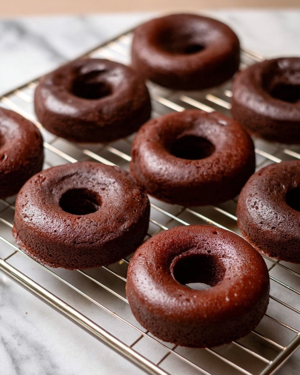 Several chocolate donuts are arranged on a metal cooling rack over a white marbled surface. Each donut has a thick, shiny layer of dark chocolate glaze on top, sprinkled with small, black chocolate sprinkles. The sides of the donuts are covered with a light dusting of granulated sugar, giving a slightly rough texture. One donut is partially eaten, showing a soft, rich, chocolatey inside. A small piece of another bitten donut lies near the bottom left corner. photo taken with an iphone --ar 4:5 --v 7