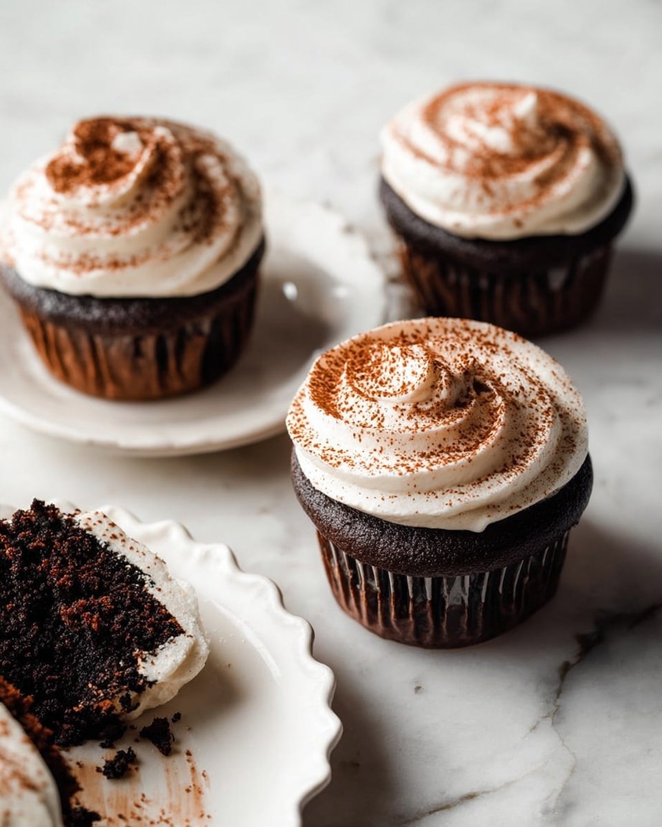 The image shows three chocolate cupcakes on a white marbled surface. Each cupcake has one dark brown chocolate cake layer, topped with a thick layer of white creamy frosting swirled in a circular motion, dusted with a fine layer of brown cocoa powder on top. Two whole cupcakes sit side by side with the frosting visible, while one cupcake is broken in pieces on a white plate with scalloped edges in the lower left corner, showing the dark chocolate crumb inside alongside the white frosting. Photo taken with an iphone --ar 4:5 --v 7