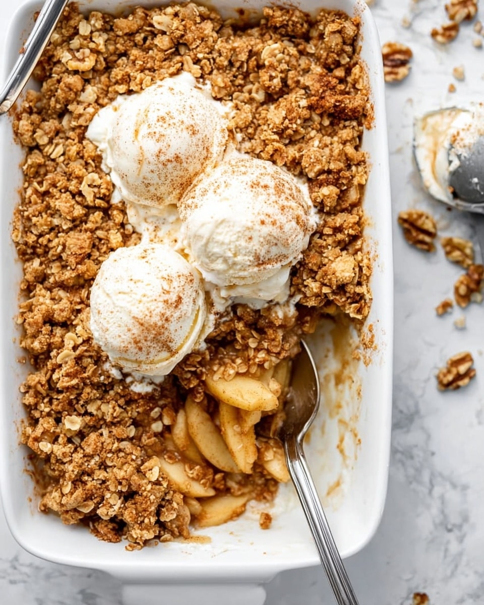 The image shows a white bowl filled with a layered apple crumble dessert. The bottom layer consists of soft, light golden-brown cooked apple slices. On top of the apples is a thick layer of golden, crunchy oat and nut crumble. Centered on the crumble is a dollop of white whipped cream sprinkled with small bits of crumble and light brown cinnamon powder. A vintage silver spoon with a textured handle rests inside the bowl, partly buried in the crumble. The bowl sits on a white marbled surface scattered with walnut pieces and cinnamon sticks. Nearby is a small white bowl with honey and a wooden honey dipper. Photo taken with an iphone --ar 4:5 --v 7
