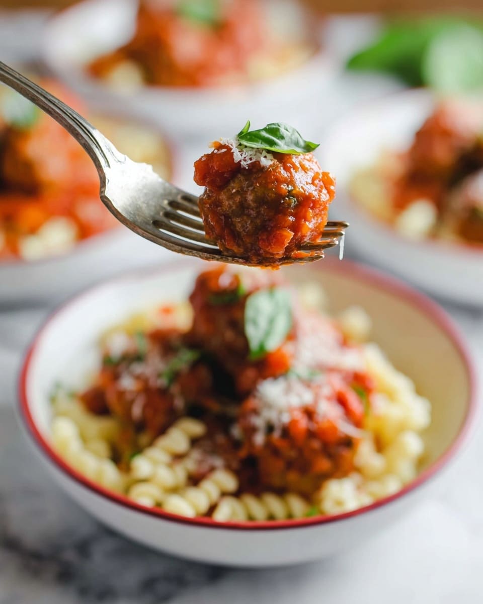 A close-up of a fork holding a meatball covered in red tomato sauce with a small green basil leaf on top, above a white bowl with a thin red rim filled with two layers: the bottom layer is pale curly pasta, and the top layer is several browned meatballs covered in chunky red tomato sauce and sprinkled with grated white cheese and a few green basil leaves; the background shows blurred white bowls with the same dish on a white marbled surface. Photo taken with an iphone --ar 4:5 --v 7